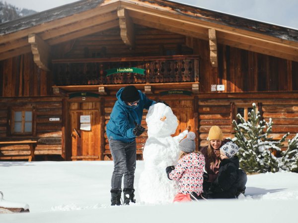 Familie baut einen Schneemann vor einer verschneiten Hütte